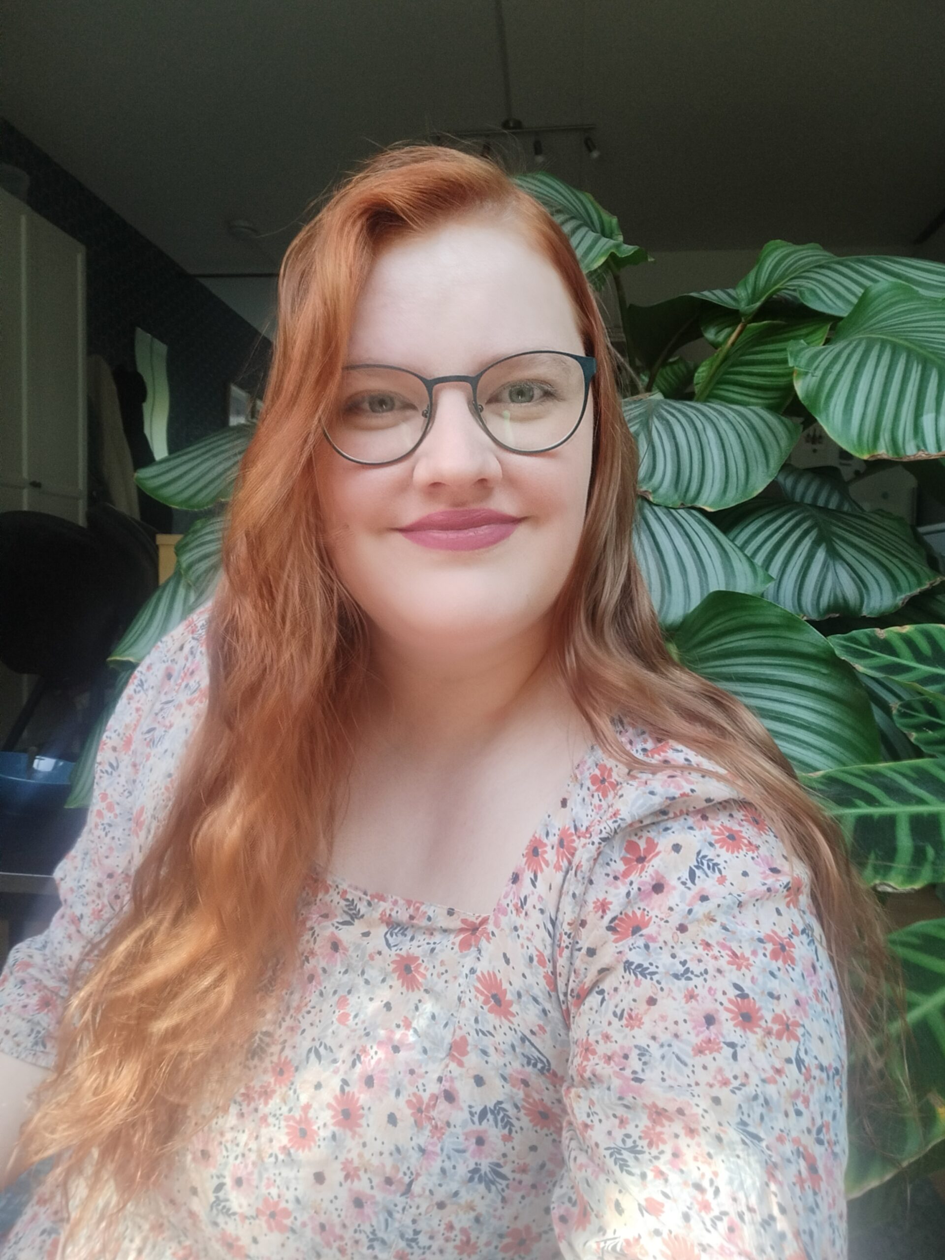IMG20220713095300 Afbeelding van Anouk, een blanke vrouw met rood lang haar, ze heeft een bloemetjes jurk aan en is geposeerd voor een groene kamerplant.